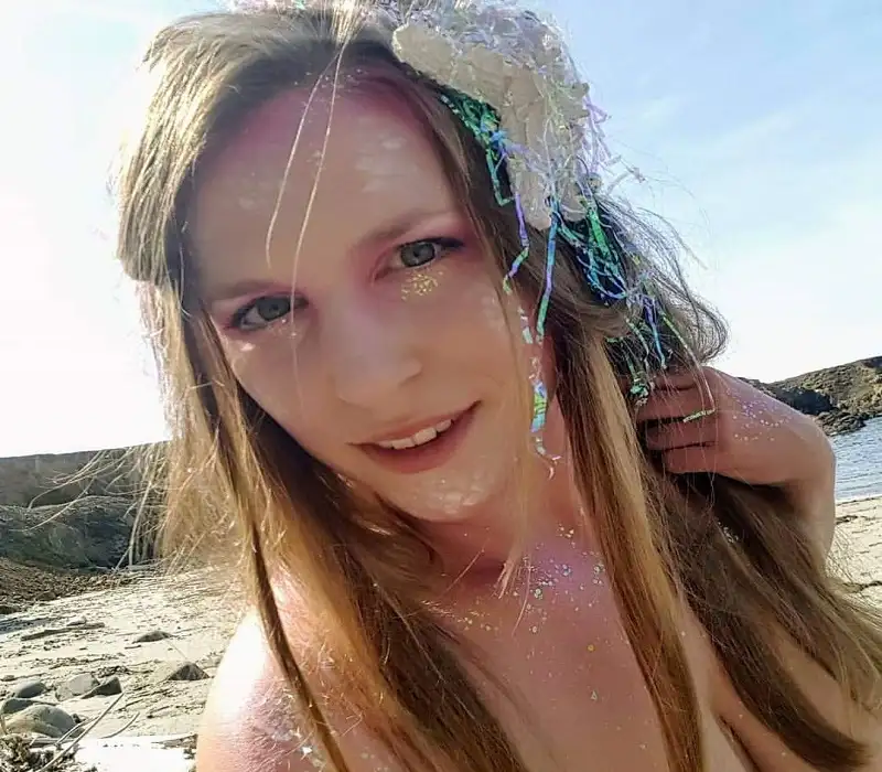 Playful woman with shimmering, ocean-inspired face makeup and flowing hair, posing on a sunny beach with rocky coastline, capturing a magical and ethereal seaside moment