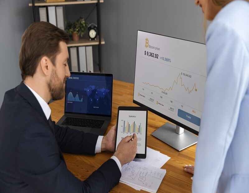 Business professionals analyzing financial graphs and cryptocurrency data on a tablet and monitor in a modern office setting.