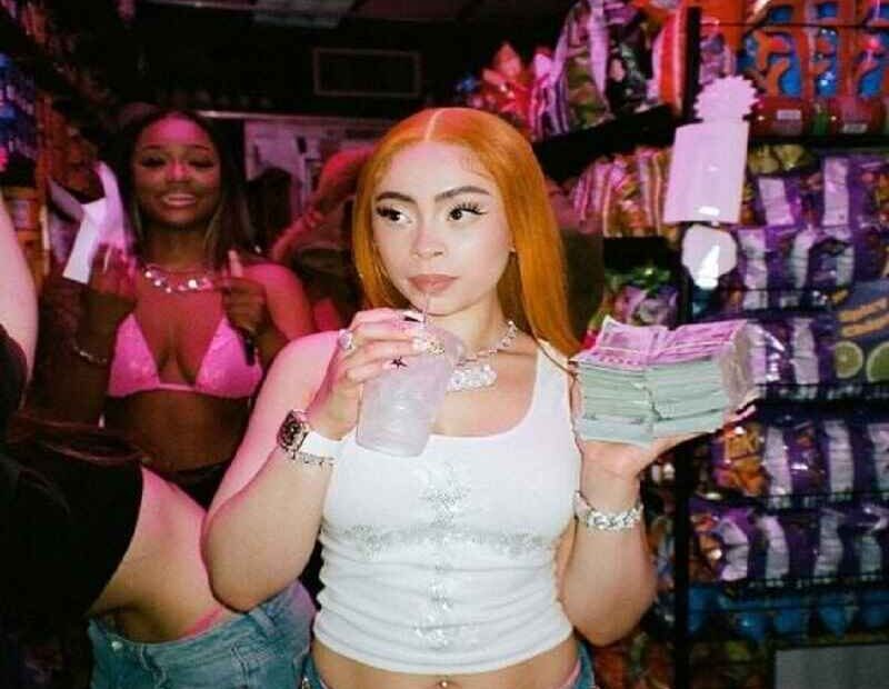 Ice Spice holding a stack of cash and sipping a drink in a vibrant store setting, showcasing her confident style.