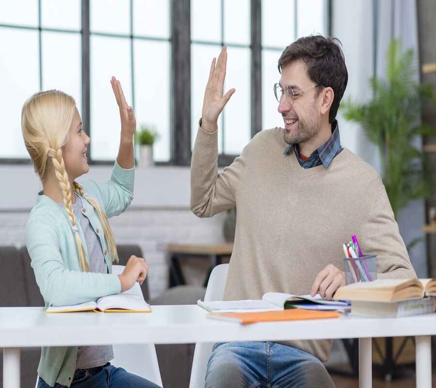 A young girl and her male tutor high-five over a study table, both smiling, with books and notebooks around them in a bright classroom setting.