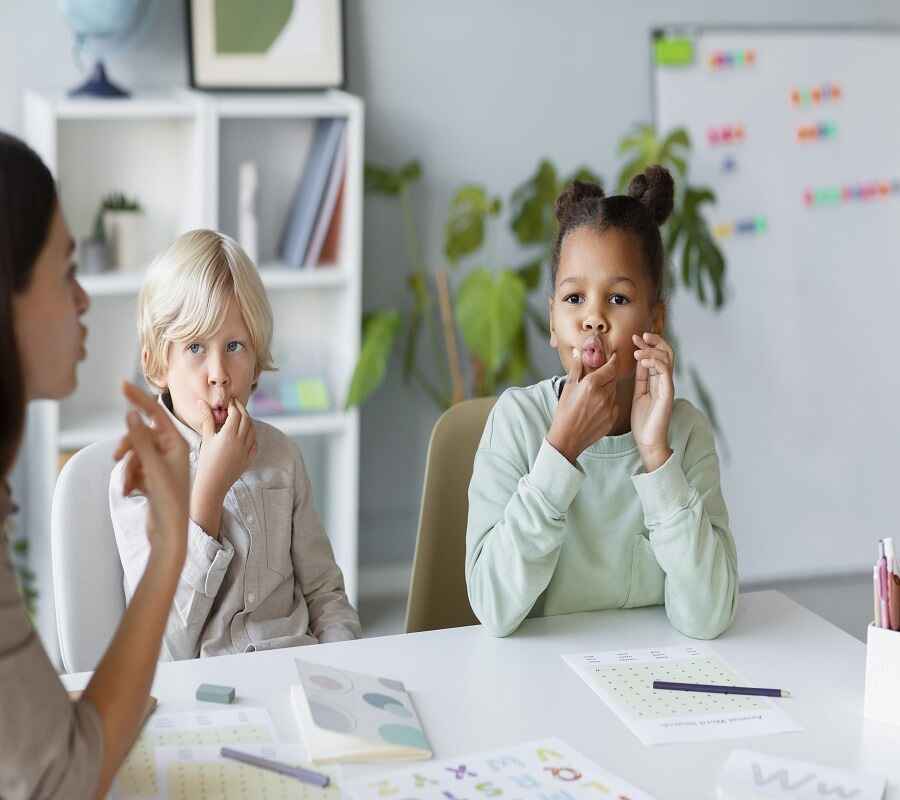 Two children in a classroom learning sign language, with a young girl instructing as the boy and another girl attentively practice the signs.