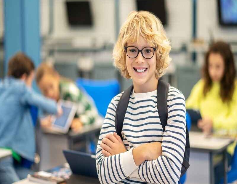 A young boy with curly blond hair and glasses, smiling in a school classroom while holding his backpack, dressed in a striped t-shirt.