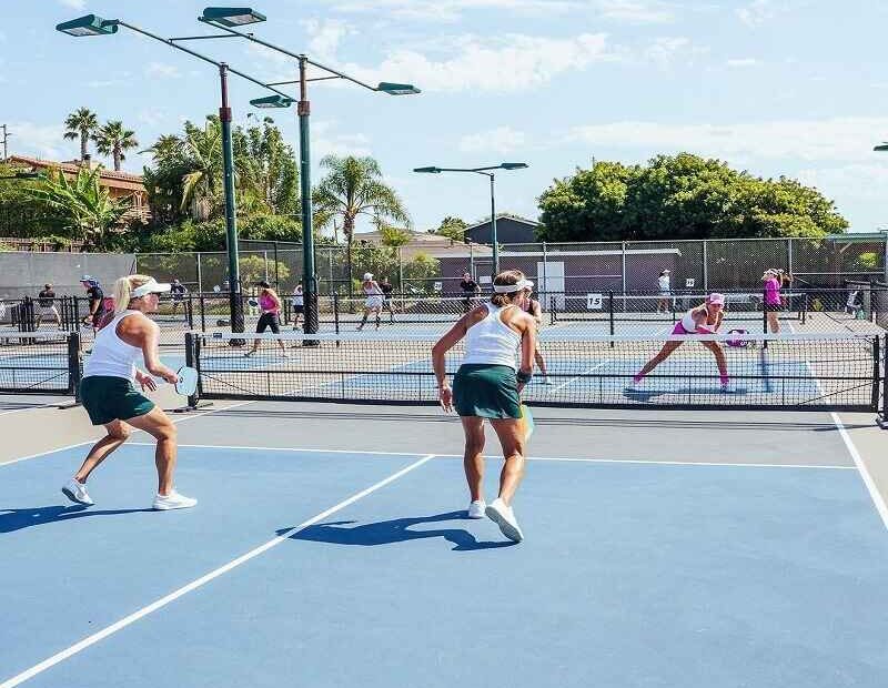 Two players on a pickleball court engaged in a match, while others play on nearby courts in a sunny outdoor setting.