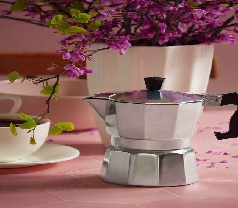 Still life setup featuring a moka pot coffee maker with a blooming plant in the background, next to a cup of coffee on a pink surface, highlighting a cozy and vibrant coffee preparation scene.