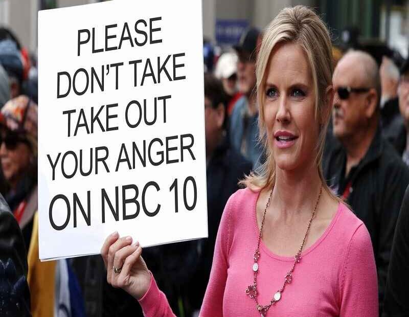 A woman in a pink sweater holds a sign that reads, "Please don't take out your anger on NBC 10," standing among a crowd. She has a calm expression, urging for peace and understanding amidst public frustration.