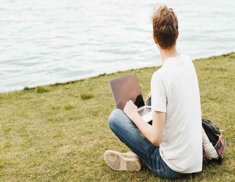 A person sitting on grass near a body of water, working on a laptop and observing the surroundings.