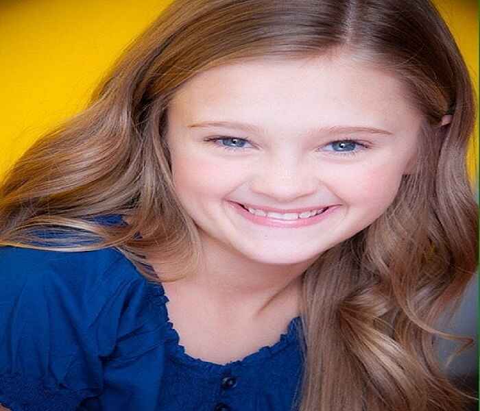 Young Lizzy Greene is smiling brightly in this close-up photo. She is wearing a blue top and has long, wavy hair. The background is yellow, highlighting her cheerful expression and blue eyes.