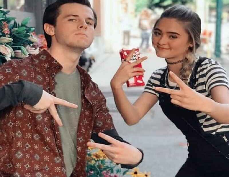 Lizzy Greene is posing with a male friend. Both are making peace signs with relaxed expressions, capturing a casual and fun moment outdoors.