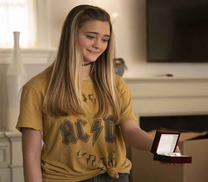 Lizzy Greene is holding an open jewelry box and smiling softly, wearing a yellow graphic t-shirt. The scene captures a thoughtful moment indoors.