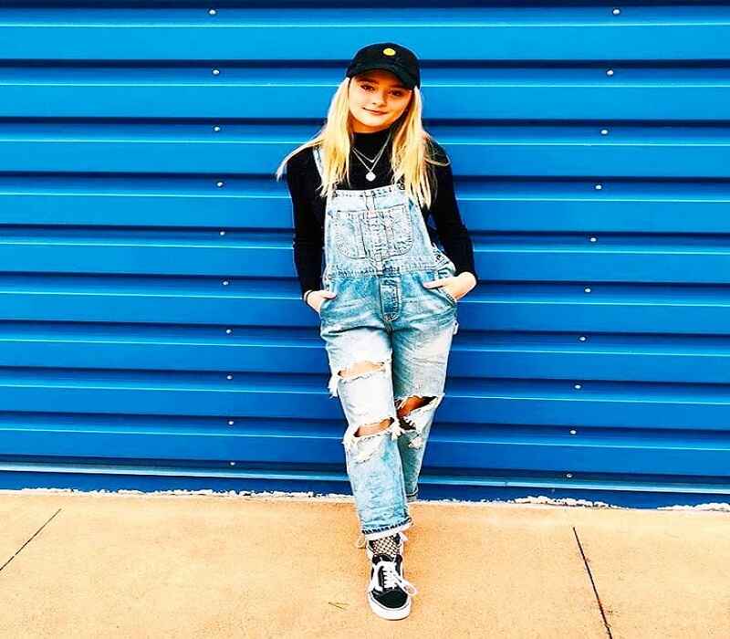 Lizzy Greene is standing in front of a blue wall, wearing ripped denim overalls, a black top, a cap, and sneakers. Her hands are in her pockets, giving off a relaxed and casual vibe.