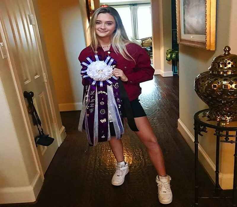 Lizzy Greene is holding a large purple ribbon while smiling indoors. She is dressed casually in a red jacket and shorts, standing confidently with one leg forward, showing her achievement with pride.