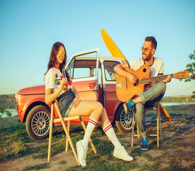 Man playing guitar while a woman sits holding a drink, both seated near a vintage car with a surfboard, enjoying an outdoor setting.