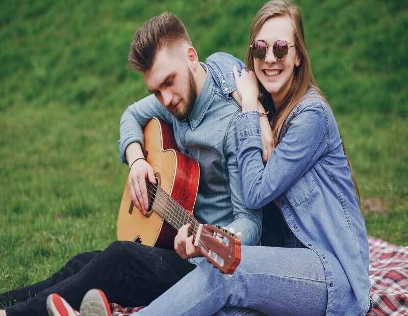 Young couple sitting outdoors on a blanket, the man playing a guitar while the woman smiles and embraces him.