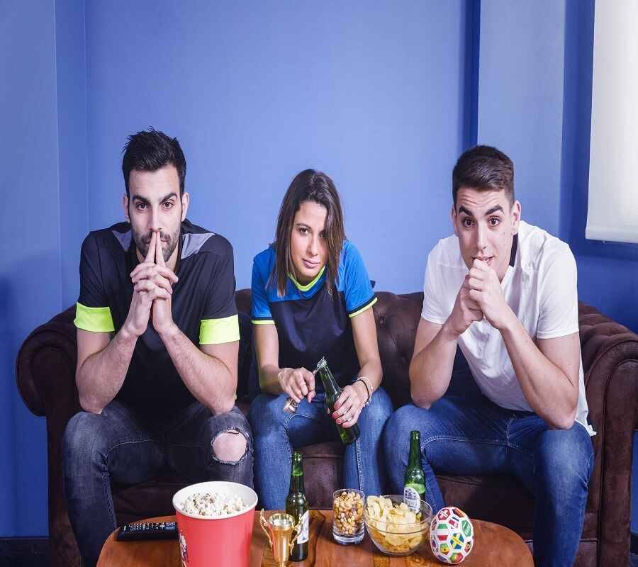 Three friends sitting on a couch, intensely watching TV, with snacks and drinks on the table.