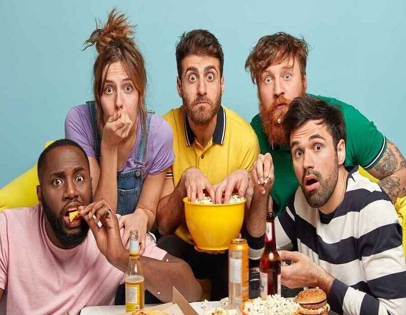 A group of five friends with animated expressions watching a movie, eating snacks like popcorn and pizza, in a lively and casual setting.