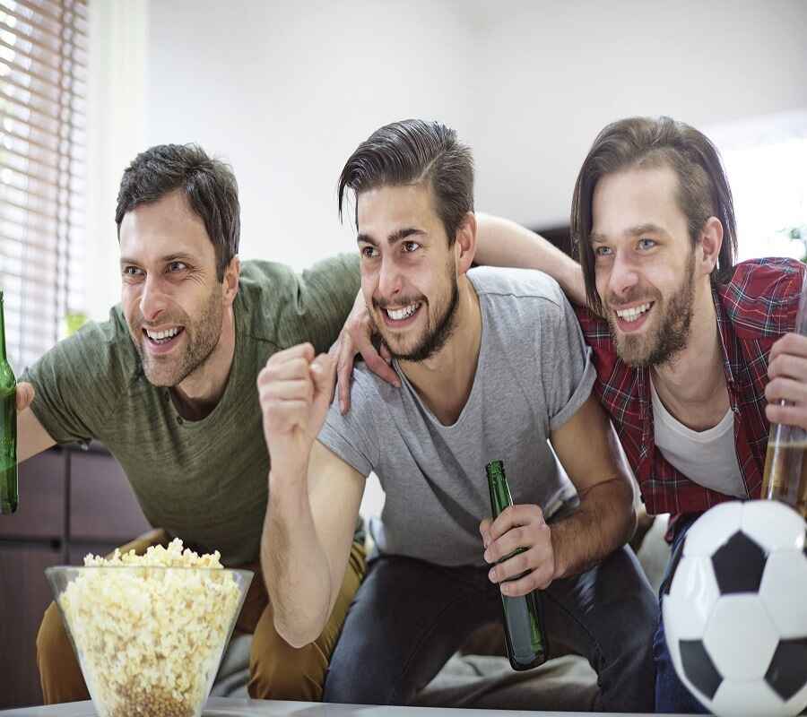 Three men cheering while watching a soccer game on TV, with beers and popcorn on the table.