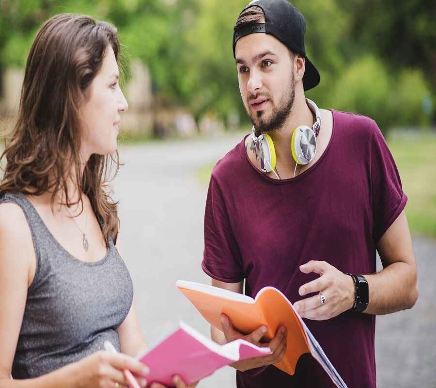 A young man with headphones around his neck discussing notes with a woman holding a book outdoors.