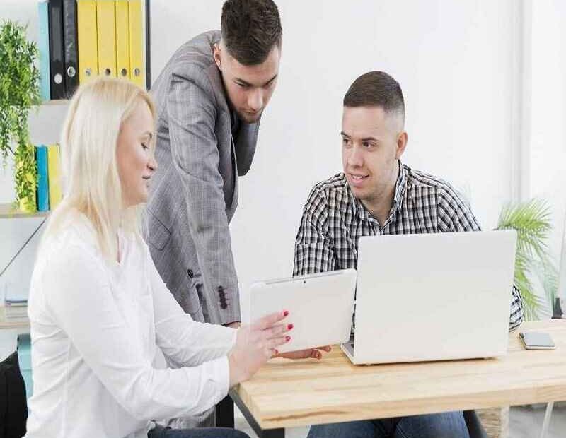 Two men and a woman in a modern office setting, reviewing data on a tablet and laptop.