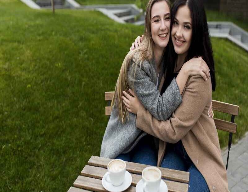 Two women sitting at an outdoor café table, smiling and hugging each other warmly while enjoying coffee together, reflecting shared joy and connection.