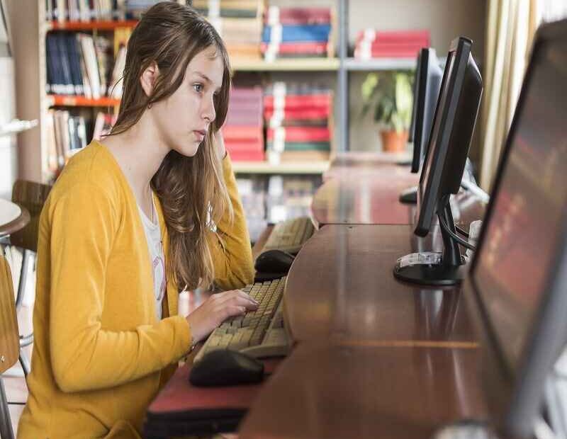 A young woman working on a computer in a library, surrounded by bookshelves, focusing intently on the screen.