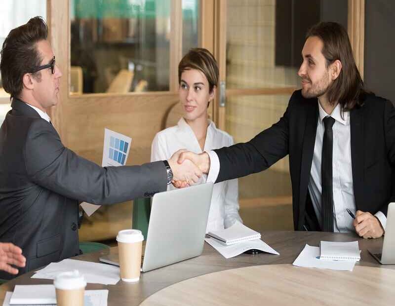 "Business professionals in a meeting room, shaking hands to finalize an agreement, while a colleague observes. A Deal OECD JanuaryLoveJoy9to5Mac