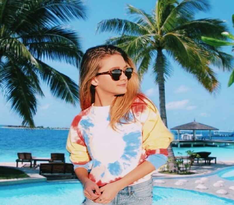 Lizzy Greene is standing outdoors by a pool, wearing sunglasses and a tie-dye sweatshirt. She is looking off to the side, with palm trees and clear blue water in the background, creating a relaxed tropical vibe.